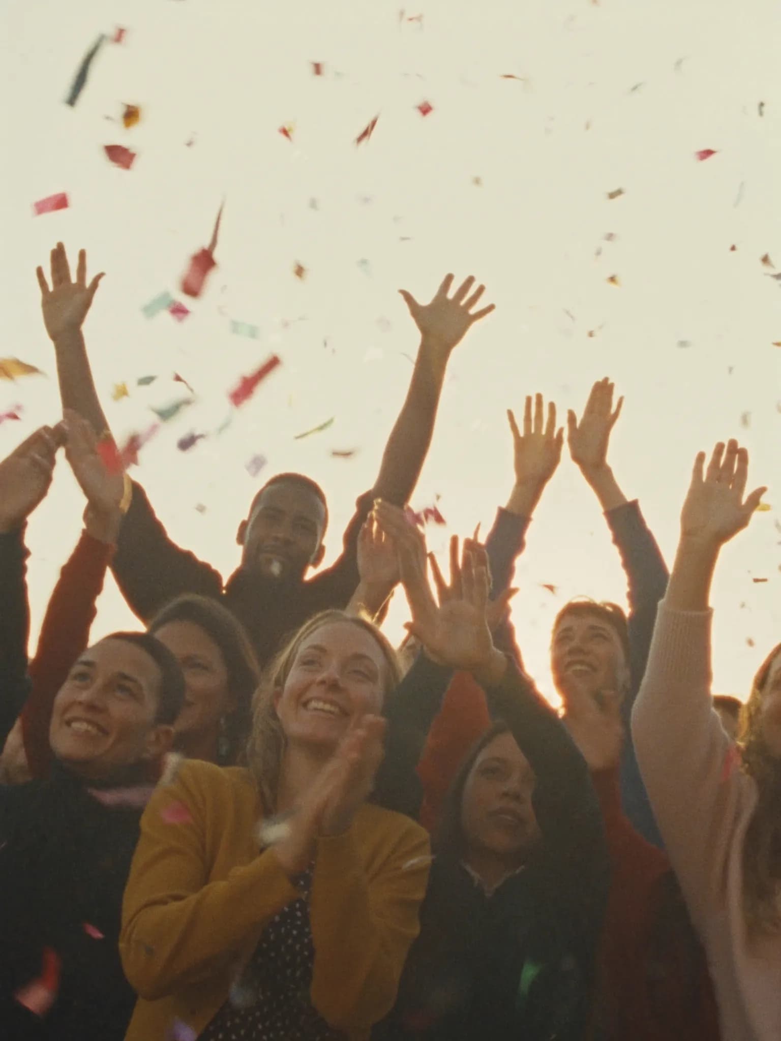 Joyful daytime rally crowd raising open hands in warm sunlight with confetti
